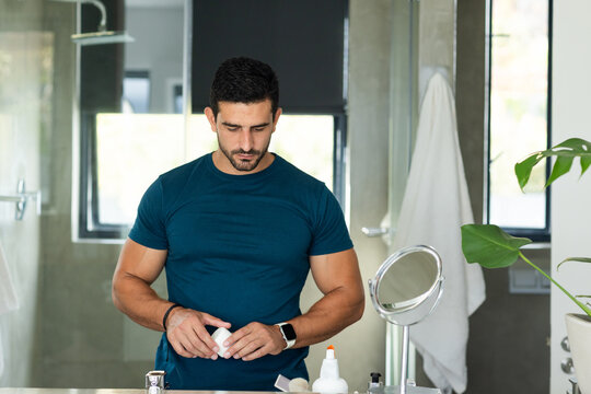 Man preparing small round grooming container at bathroom sink with mirror, pump bottle, plant