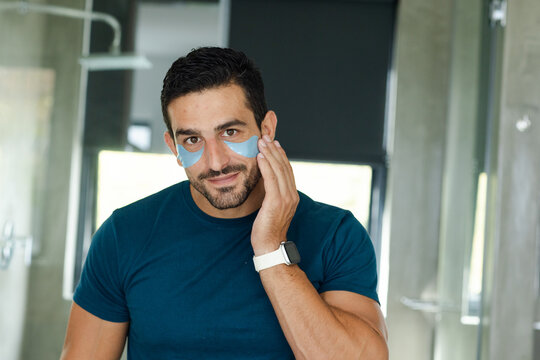 Man in navy tee adjusting blue eye patches while checking bathroom mirror, white smartwatch