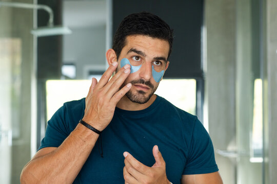 Man applying under-eye gel patches while looking into mirror in modern bathroom wearing teal shirt