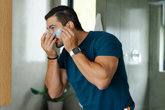 Adult man applying blue under-eye gel patch at bathroom sink checking mirror, wearing smartwatch