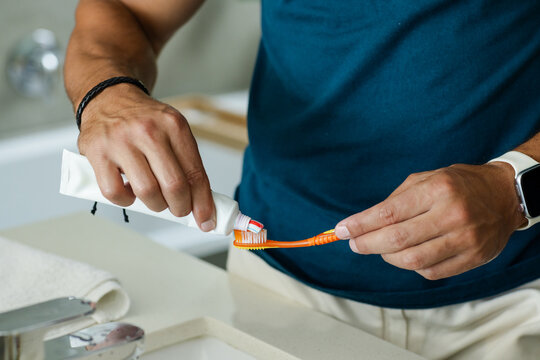 Adult man squeezing toothpaste onto orange toothbrush at bathroom sink, wearing watch and band