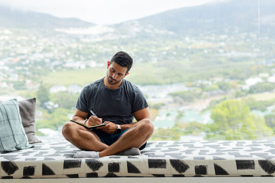 Man sitting cross-legged on patterned bench by floor-to-ceiling window, writing in notebook