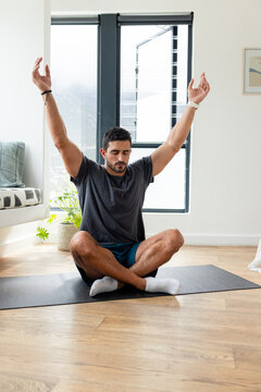 Adult man sitting cross-legged on dark exercise mat in bright living room, wearing sportswear