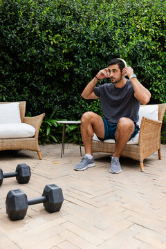 Adult man adjusting white headphones while sitting on paved patio with hexagonal dumbbells nearby