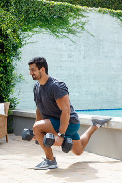 Man doing single-leg lunge on patio holding dumbbells using low wall wearing shorts, smartwatch