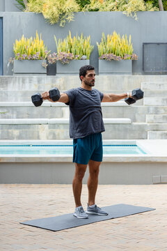 Man in his 30s performing lateral raises on dark mat beside pool holding hex dumbbells