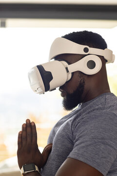 African man standing at home near window wearing white VR headset and grey tee pressing palms