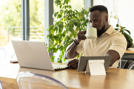 Man wearing polo sitting at home table working on laptop while holding mug and tablet