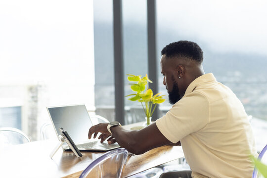 Man sitting at high wooden table, wearing polo and wristwatch, using laptop and tablet, copy space