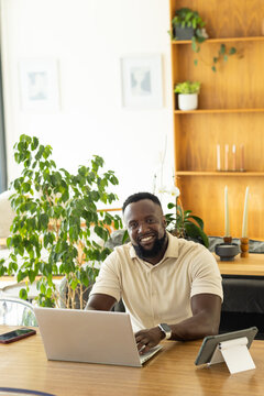 African adult man sitting at home office working on silver laptop wearing beige polo and smartwatch
