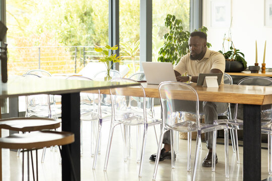 African American man in polo-shirt working on laptop at wooden-table with clear chairs, copy space