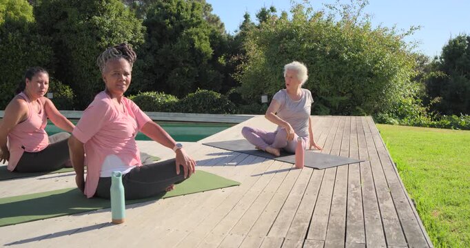 Female friends, senior doing yoga seated twists on deck with mats and bottles as pillar clearing
