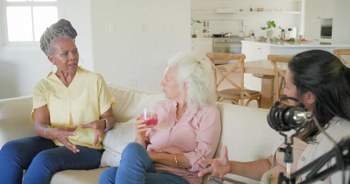 Diverse women on sofa, host asking with tablet to record near mic, guests gesturing, sipping glass