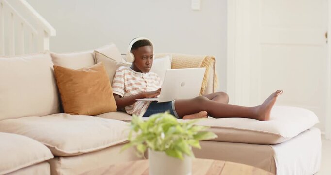 African youth male settling into couch, typing on laptop, wearing headphones and maintaining focus