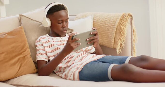 African male teen leaning back on sofa at home, tapping tablet and browsing content wearing headset