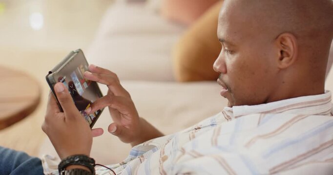 African American man on sofa holding tablet with earphones, tapping, swiping tiles, selecting tile