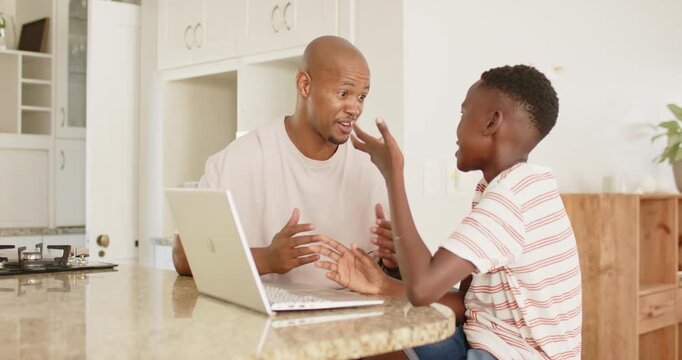 African father and boy sitting at kitchen counter, father pointing at laptop, son typing to learn