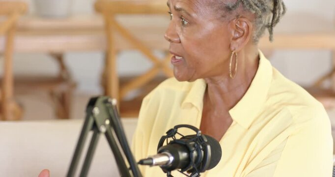 Senior woman is lifting metal cup to offer condiment tasting while speaking into mic on sofa