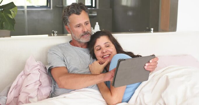 Mature couple cuddling in bed, man holding tablet browsing content, woman guiding taps, relaxing