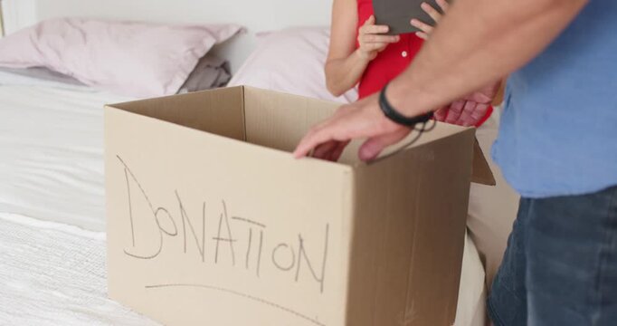Couple packing donation box on bed, woman looking up and checking tablet while man folding flaps
