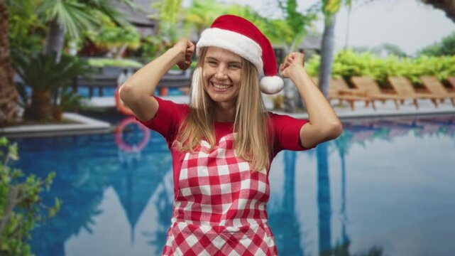 Woman raises arms wearing santa hat and red gingham apron beside a resort pool inside a building, smiling and dancing; holiday joy.