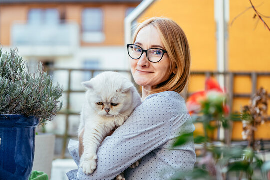 Warm middle aged caucasian woman holding white cat on balcony looking at camera