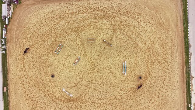 Horses move around an outdoor riding arena with several jumps placed on the sandy surface. The scene shows clear markings from horses and obstacles in a circular pattern.