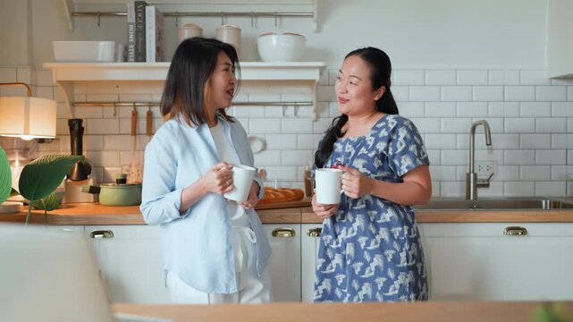 Two Asian women friends standing in kitchen talking and laughing together, enjoying good old day memories, relaxed casual moment, warm home lifestyle, friendship bonding and cheerful social connection