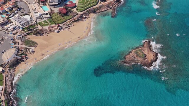 Aerial drone view of Fig Tree Bay, Protaras, Cyprus, with its iconic small offshore islet surrounded by crystal clear water and a sandy beach lined with hotels