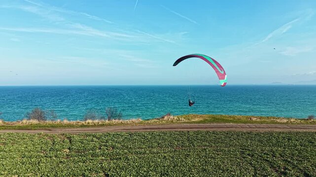 A person is paragliding above the ocean while the sun shines. Below, there are fields of green along the coast. The sky is blue, and the water sparkles in the sunlight.
