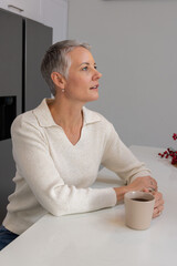 Senior woman sitting at kitchen counter holding beige mug on countertop in cream sweater, looking © wavebreak3