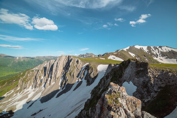 Aerial dizzying view to rocky cliff among sharp rocks under clouds in blue sky. Vertigo scenery with sheer crags above precipice, snowy ridge and mountain top far away in cloudy changeable weather. © Daniil