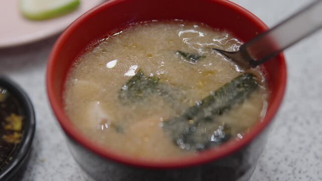 Spoon gently stirs the rich broth of a red and black bowl of traditional japanese miso soup, containing soft tofu and fresh seaweed, highlighting a moment of culinary enjoyment