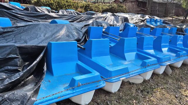 Row of blue pedal boats stored on shore and covered with protective tarps, early spring outdoor scene with dry grass and soft sunlight, concept of seasonal reopening and recreation