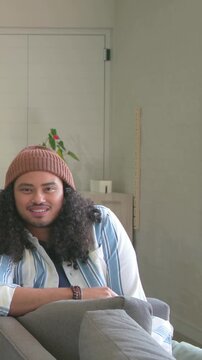 Vertical video: Straightening man wearing beanie adjusting cushion on sofa, reacting to left person