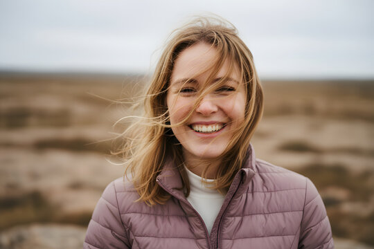 Laughing blonde woman outdoors with windblown hair, wearing a pink puffer jacket, smiling at the camera in a rugged natural landscape under soft overcast light.