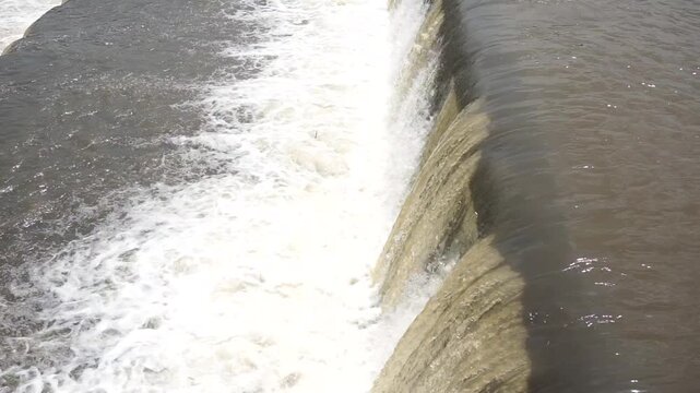 the flow of river water that passes over a small dam or threshold, creating a waterfall effect with white foam that contrasts with the brownish color of the river water
