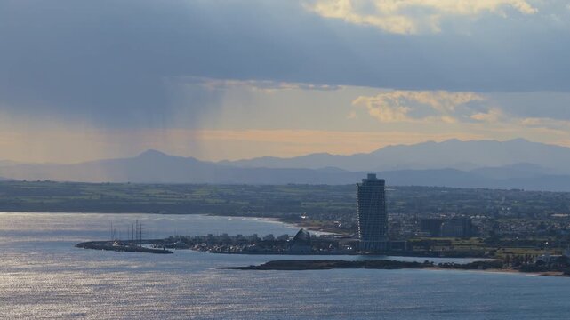 Aerial drone view of coastline featuring a tall beachfront hotel and sun rays breaking through clouds over the Mediterranean Sea