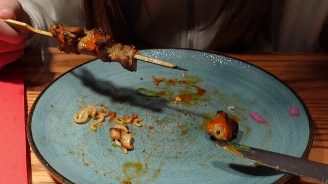 A grilled meat skewer served over couscous with vibrant orange sauce and microgreens, served in rustic bowl on wooden table of a restaurant and woman eating in a second dish
