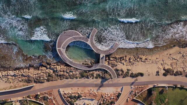 Aerial drone view of a unique curved wooden pier over the sea in Ayia Napa, Cyprus, surrounded by waves and rocky coastline