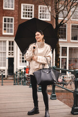Obraz premium A joyful woman in stylish beige trench coat, black leggings, and chunky boots stands on a wooden bridge holding an open black umbrella with a wide smile in an urban setting with historic architecture