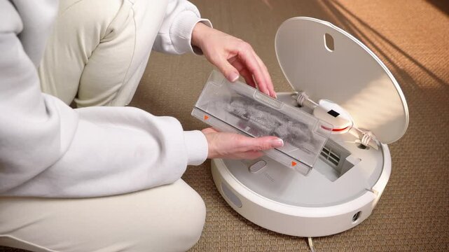 Woman maintaining robotic vacuum cleaner dust bin filter. Close-up of female hands removing a full dustbin container from a robotic vacuum cleaner to clean the dirty filter