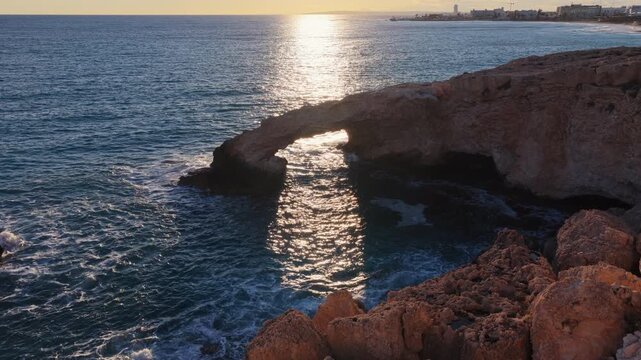 Aerial drone view of the Bridge of Love, a natural rock arch formation in Ayia Napa, Cyprus. Turquoise waves flow beneath the limestone arch