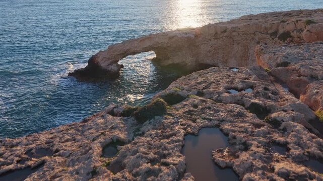 Aerial drone view of the Bridge of Love, a natural rock arch formation in Ayia Napa, Cyprus. Turquoise waves flow beneath the limestone arch