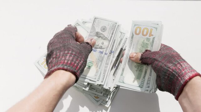 Adult beggar counts one hundred dollar bills indoors against a plain white background.