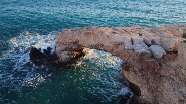 Aerial drone view of the Bridge of Love, a natural rock arch formation in Ayia Napa, Cyprus. Turquoise waves flow beneath the limestone arch
