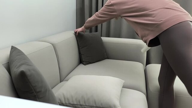 Hand uses lint roller cleaning velour pillow on sofa at home. Person removes hair and dust from upholstery surface indoors.