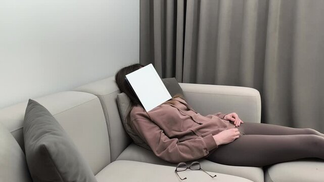 Woman falls asleep on sofa with book covering face at evening. Female rests after reading placing object over head indoors. Fatigue relaxation and bedtime concept in apartment living room..