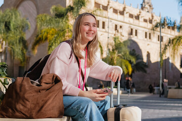 Fototapeta premium Solo female traveler with rolling suitcase checks smartphone navigation in sunny European city, city break weekend getaway, urban tourism, travel planning app, digital nomad lifestyle.
