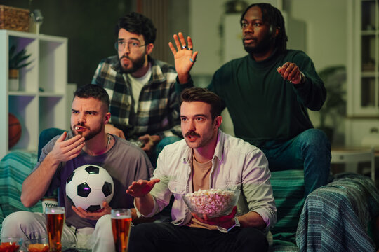 Diverse male friends watching exciting soccer match on tv, celebrating victory and sharing snacks at home